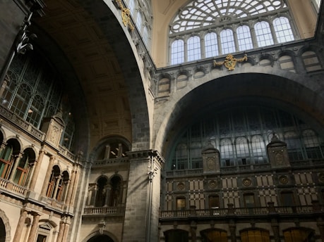 The image captures an elegant architectural interior featuring large, arched windows with intricate metal and stone detailing. The walls are adorned with decorative elements and columns, showcasing a blend of classical and industrial styles.