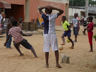 Children are playing soccer on a sandy ground in an urban neighborhood. Various buildings and structures are visible in the background, as well as a few parked vehicles. The kids are in motion, engaged in the game, with one child about to kick a ball.