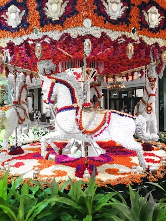A vibrant carousel decorated entirely with colorful flowers. Intricate patterns of orange, red, purple, and white flowers create the shape of carousel horses surrounded by floral arrangements. Lanterns hang around the structure, adding a decorative touch.