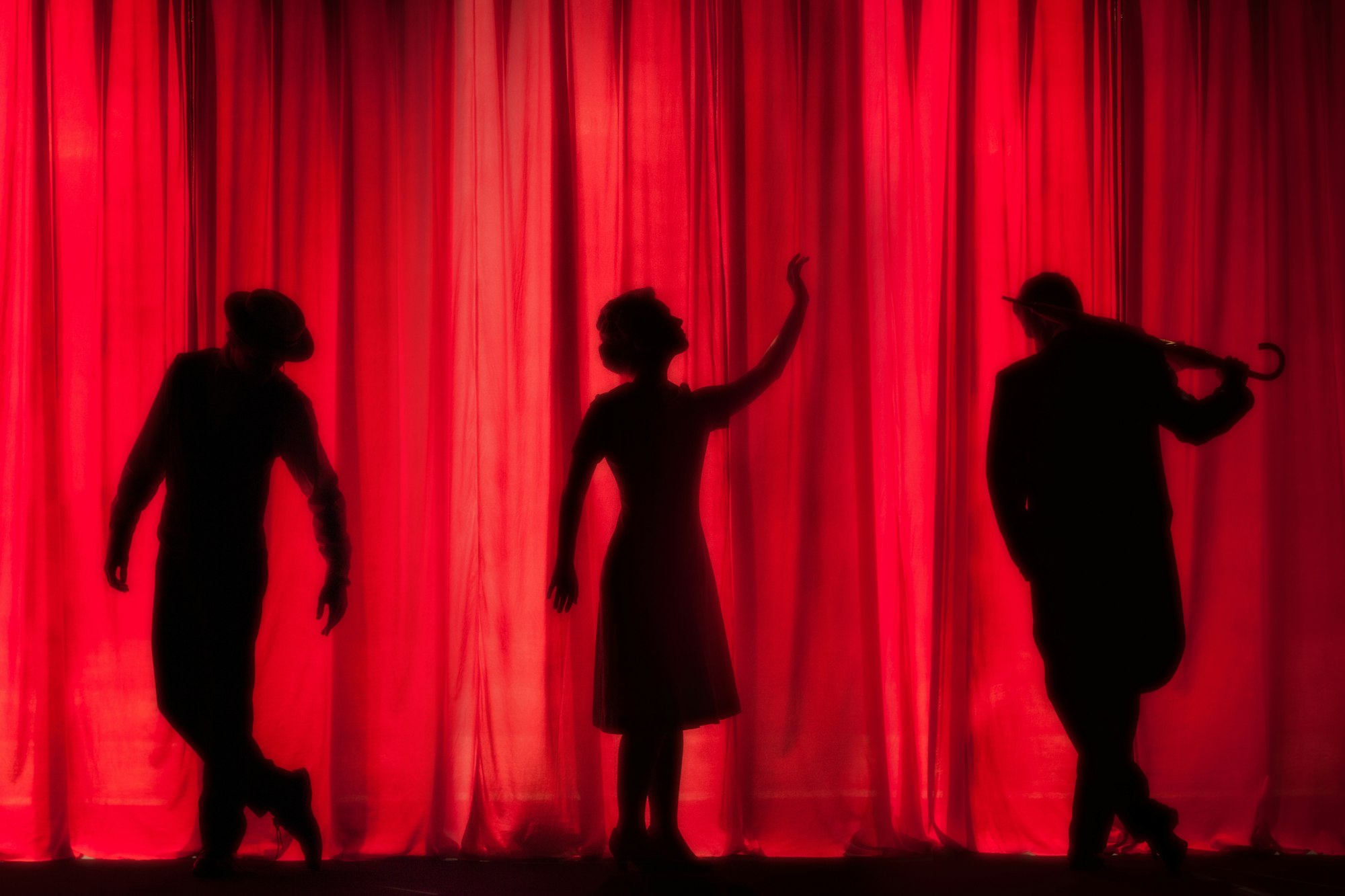 The silhouettes of 3 people acting in front of a red curtain