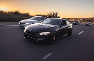 A group of sleek, modern cars driving swiftly down a highway at sunset, with the sky displaying warm hues. The foreground features a shiny black car, prominently displayed, followed by a silver car to its side. The road is wide and mostly empty, lined by blurred green foliage indicating motion.