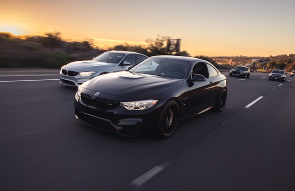 A group of sleek, modern cars driving swiftly down a highway at sunset, with the sky displaying warm hues. The foreground features a shiny black car, prominently displayed, followed by a silver car to its side. The road is wide and mostly empty, lined by blurred green foliage indicating motion.