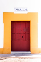 A red wooden door with black studs is framed by a vibrant yellow border set into a white wall. Above the door, a sign reads 'Taquillas' in bold blue lettering, indicating ticket booths. The bottom of the image shows a stone pavement against the wall.