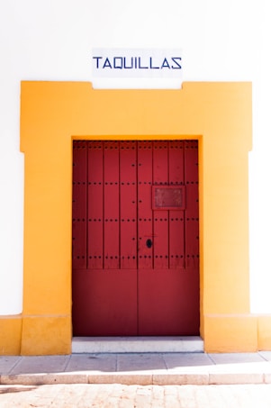 A red wooden door with black studs is framed by a vibrant yellow border set into a white wall. Above the door, a sign reads 'Taquillas' in bold blue lettering, indicating ticket booths. The bottom of the image shows a stone pavement against the wall.