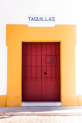 A red wooden door with black studs is framed by a vibrant yellow border set into a white wall. Above the door, a sign reads 'Taquillas' in bold blue lettering, indicating ticket booths. The bottom of the image shows a stone pavement against the wall.