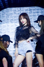 A person in a black sheer blouse and denim shorts poses confidently on stage, surrounded by dancers in black outfits and caps. The background features a textured brick wall with dramatic lighting.