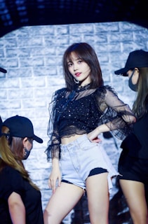 A person in a black sheer blouse and denim shorts poses confidently on stage, surrounded by dancers in black outfits and caps. The background features a textured brick wall with dramatic lighting.