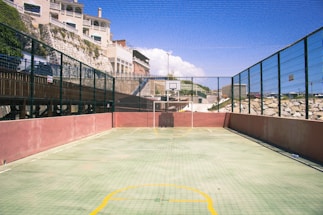 An outdoor sports court with fencing on both sides and a basketball hoop at the far end. The surface is marked with yellow lines and surrounded by a high wire mesh. There are rocks and a building in the background under a clear blue sky.