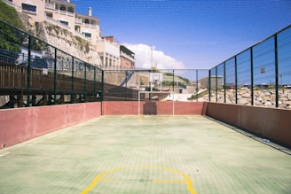 An outdoor sports court with fencing on both sides and a basketball hoop at the far end. The surface is marked with yellow lines and surrounded by a high wire mesh. There are rocks and a building in the background under a clear blue sky.