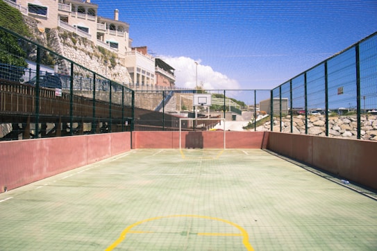 An outdoor sports court with fencing on both sides and a basketball hoop at the far end. The surface is marked with yellow lines and surrounded by a high wire mesh. There are rocks and a building in the background under a clear blue sky.