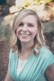 A blonde-haired woman with a bright smile is outdoors, wearing a light teal top over a white shirt. She has wavy hair and is positioned in a slightly shaded environment, with rocks and some greenery in the blurred background.