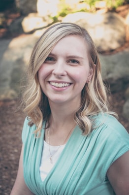 A blonde-haired woman with a bright smile is outdoors, wearing a light teal top over a white shirt. She has wavy hair and is positioned in a slightly shaded environment, with rocks and some greenery in the blurred background.