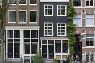 A row of traditional European townhouses with brick facades and large windows. The central building has a dark exterior with ivy climbing up one side. People are casually riding bicycles in the foreground, creating a lively urban atmosphere.