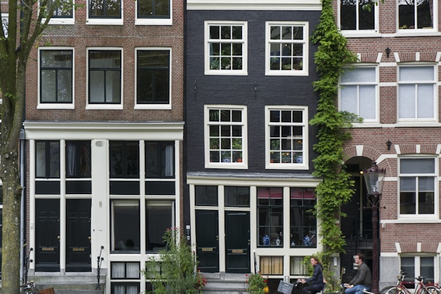 A row of traditional European townhouses with brick facades and large windows. The central building has a dark exterior with ivy climbing up one side. People are casually riding bicycles in the foreground, creating a lively urban atmosphere.