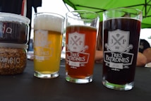 Three glasses of beer in differing colors are placed on a table, each with the Tres Patrones logo printed on them. The beers vary from pale yellow to amber to dark brown. Next to the beers, there are containers filled with what appears to be grains and another with a dark substance, likely for brewing. A green canopy is visible above, suggesting an outdoor setting.