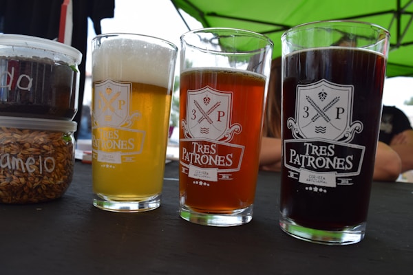 Three glasses of beer in differing colors are placed on a table, each with the Tres Patrones logo printed on them. The beers vary from pale yellow to amber to dark brown. Next to the beers, there are containers filled with what appears to be grains and another with a dark substance, likely for brewing. A green canopy is visible above, suggesting an outdoor setting.