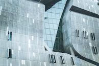 A modern architectural structure featuring a facade with angled, metallic panels and large glass windows. The surface showcases a grid pattern, with smooth, curved sections mixing with angular elements, creating a futuristic appearance.