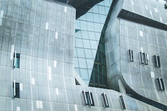 A modern architectural structure featuring a facade with angled, metallic panels and large glass windows. The surface showcases a grid pattern, with smooth, curved sections mixing with angular elements, creating a futuristic appearance.