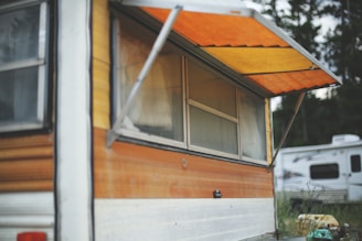 A vintage camper with a wooden exterior and an extended orange awning is in focus. The window is framed by aluminum and partially open, and behind it is greenery. Another camper is visible in the background, positioned among some plants and an outdoor setting.