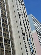 A cityscape featuring tall buildings with modern architectural designs against a clear blue sky. One of the buildings prominently displays a sign that reads 'CESAR BUSINESS'. The structures have a variety of rectangular windows, and one building has a beige and cream façade.