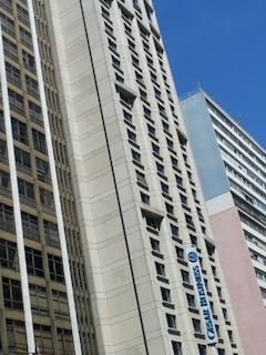 A cityscape featuring tall buildings with modern architectural designs against a clear blue sky. One of the buildings prominently displays a sign that reads 'CESAR BUSINESS'. The structures have a variety of rectangular windows, and one building has a beige and cream façade.