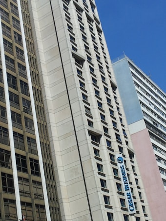 A cityscape featuring tall buildings with modern architectural designs against a clear blue sky. One of the buildings prominently displays a sign that reads 'CESAR BUSINESS'. The structures have a variety of rectangular windows, and one building has a beige and cream fa&ccedil;ade.