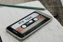 A smartphone displaying an image of a retro cassette tape labeled 'Mix Tape' is placed on an open notebook filled with handwritten notes. A pencil rests on top of the notebook, and the scene is set on a wooden surface.