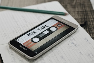 A smartphone displaying an image of a retro cassette tape labeled 'Mix Tape' is placed on an open notebook filled with handwritten notes. A pencil rests on top of the notebook, and the scene is set on a wooden surface.