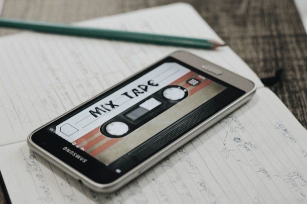 A smartphone displaying an image of a retro cassette tape labeled 'Mix Tape' is placed on an open notebook filled with handwritten notes. A pencil rests on top of the notebook, and the scene is set on a wooden surface.