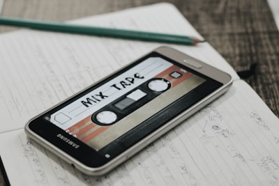 A smartphone displaying an image of a retro cassette tape labeled 'Mix Tape' is placed on an open notebook filled with handwritten notes. A pencil rests on top of the notebook, and the scene is set on a wooden surface.