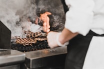 A chef grills hamburger patties on a commercial grill, surrounded by smoke and flames. The patties show distinct grill marks, and the chef is wearing a white coat and black apron, focusing on the cooking process.