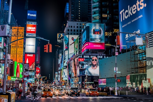 A vibrant city street landscape at night, featuring numerous illuminated billboards and signs. Bright advertisements for major brands dominate the scene, while yellow taxis and people walk along the busy street below. Tall buildings frame the backdrop, contributing to the lively urban atmosphere.