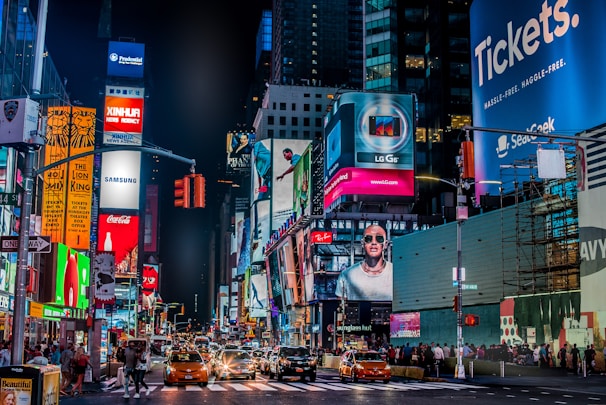 A vibrant city street landscape at night, featuring numerous illuminated billboards and signs. Bright advertisements for major brands dominate the scene, while yellow taxis and people walk along the busy street below. Tall buildings frame the backdrop, contributing to the lively urban atmosphere.