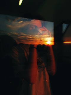 A reflective image combining a person's silhouette holding a camera with a vibrant sunset over a cloudy sky, seen through a window pane. The blending of the interior scene and the external view creates a juxtaposition of human presence and nature's beauty.