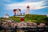 A picturesque coastal scene featuring a lighthouse with a red roof situated on a grassy hill. The lighthouse is accompanied by a traditional house with an American flag and a small white outbuilding. The area is surrounded by rocky outcrops and lush greenery under a blue sky with soft clouds.