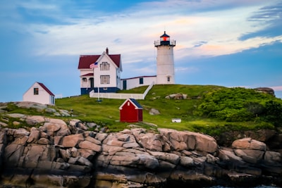 A picturesque coastal scene featuring a lighthouse with a red roof situated on a grassy hill. The lighthouse is accompanied by a traditional house with an American flag and a small white outbuilding. The area is surrounded by rocky outcrops and lush greenery under a blue sky with soft clouds.