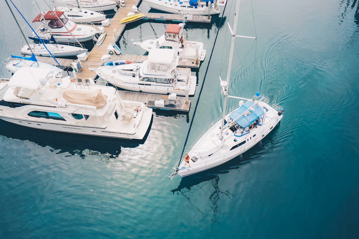 An aerial view of yachts and a sailboat berthed along a marina dock in deep teal water.