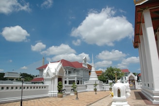 A well-maintained courtyard in a temple complex with white buildings adorned with traditional Thai architectural details and red roofs. A bright blue sky peppered with fluffy clouds creates a serene backdrop. Ornamental spires and neatly trimmed shrubs add elegance to the surroundings.