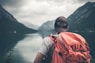 man with red hiking backpack facing body of water and mountains at daytime