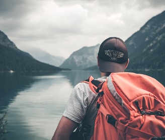 man with red hiking backpack facing body of water and mountains at daytime