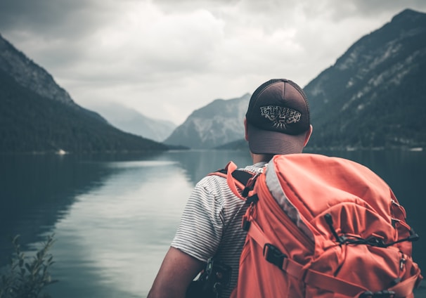 man with red hiking backpack facing body of water and mountains at daytime