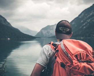 man with red hiking backpack facing body of water and mountains at daytime