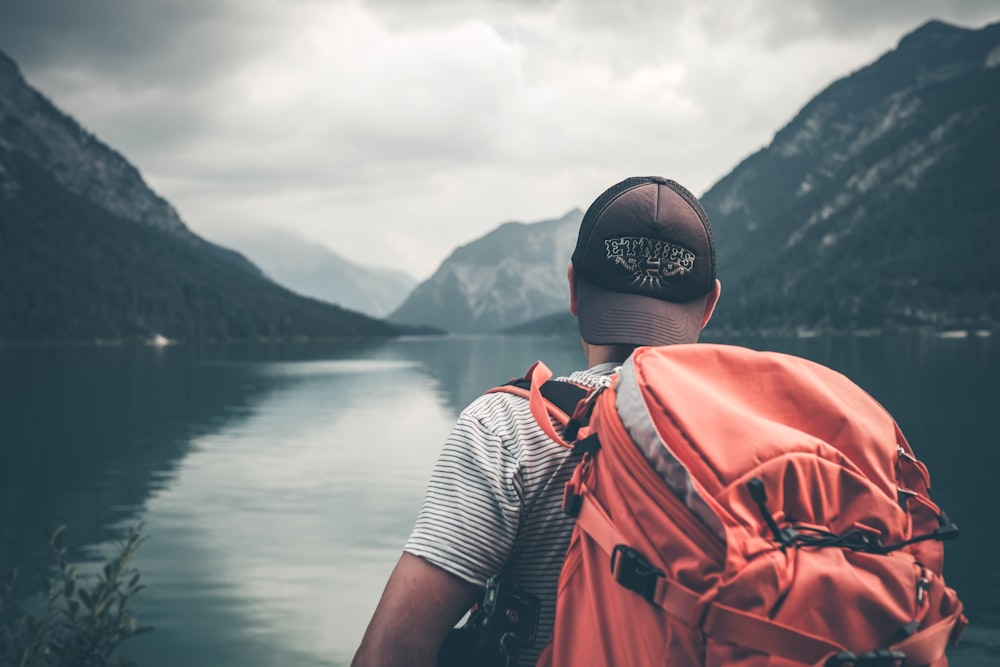 昼間、水域と山に面した赤いハイキングバックパックを背負った男性