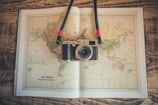 A vintage-style camera rests on an open world map book placed on a wooden surface. The map depicts a Mercator projection and has earthy tones. The camera has a strap with red accents.