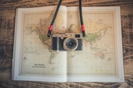 A vintage-style camera rests on an open world map book placed on a wooden surface. The map depicts a Mercator projection and has earthy tones. The camera has a strap with red accents.
