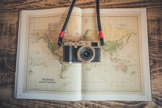 A vintage-style camera rests on an open world map book placed on a wooden surface. The map depicts a Mercator projection and has earthy tones. The camera has a strap with red accents.