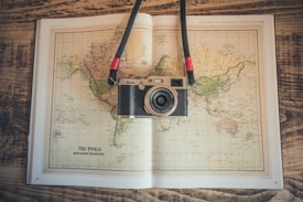 A vintage-style camera rests on an open world map book placed on a wooden surface. The map depicts a Mercator projection and has earthy tones. The camera has a strap with red accents.