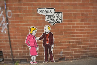 Street art on a brick wall depicting two stylized cartoon-like figures. One is a woman in a pink outfit, and the other is a man in a suit holding a speech bubble with the words 'Sydney, we need to talk!!'.