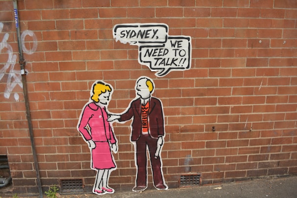 Street art on a brick wall depicting two stylized cartoon-like figures. One is a woman in a pink outfit, and the other is a man in a suit holding a speech bubble with the words 'Sydney, we need to talk!!'.