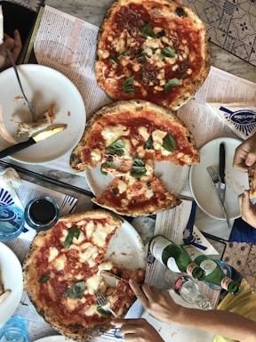 Three pizzas with tomato sauce, mozzarella cheese, and basil leaves are placed on a table. Some slices have been removed, and a couple of hands are reaching for slices. Next to the pizzas are sparkling water bottles and a glass. Plates and cutlery are scattered around.